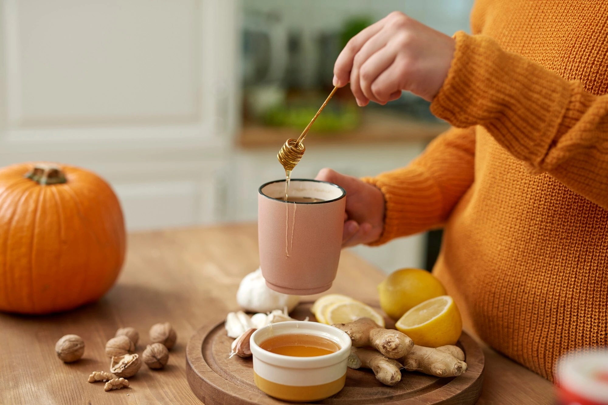 A person drizzling honey into a mug beside lemon, ginger, and pumpkin