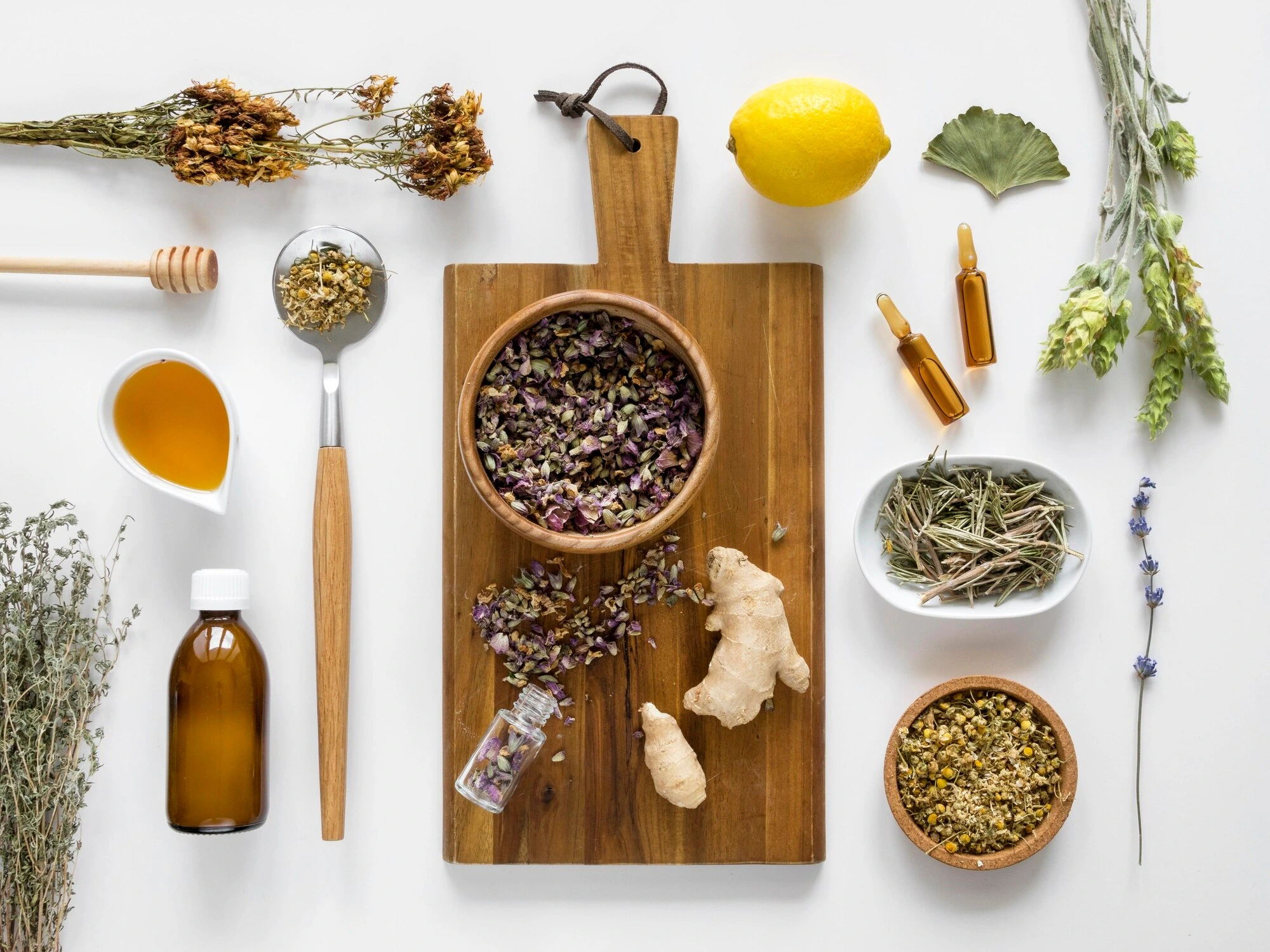 An overhead arrangement of dried herbs, tinctures, honey, lemon and ginger on a white surface