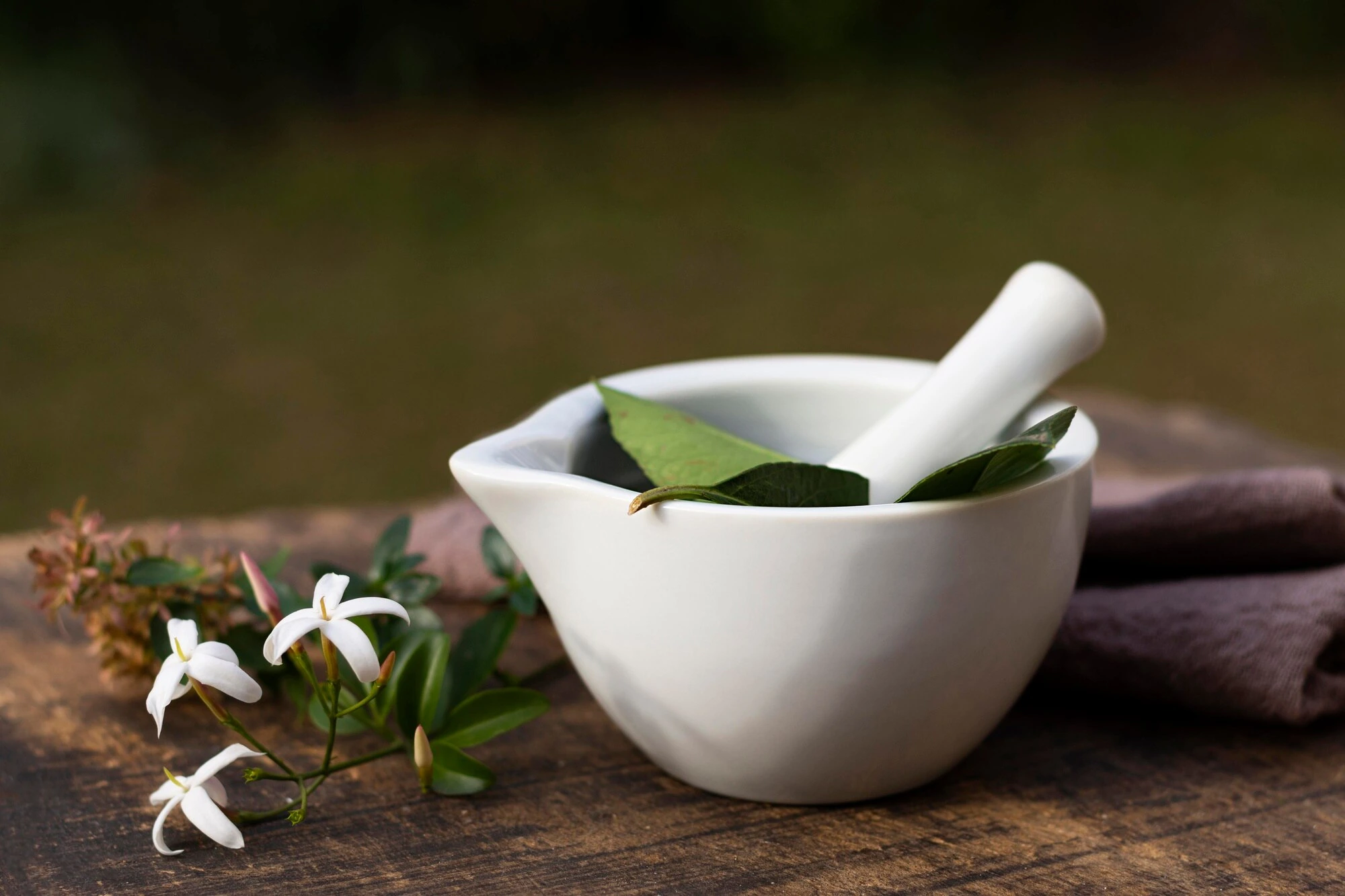 A mortar and pestle with fresh green leaves beside jasmine flowers on a wooden surface